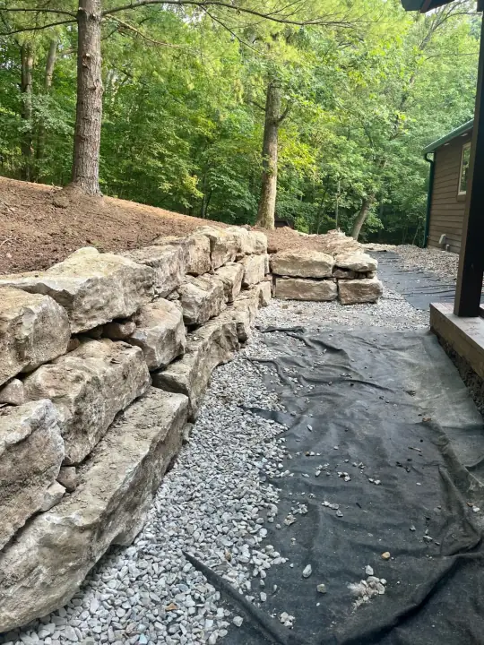A stacked boulder retaining wall beside a cabin, with black fabric and gravel along its base