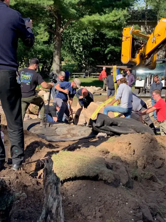 A crew working in the soil beside a stone water feature, with a yellow machine behind them