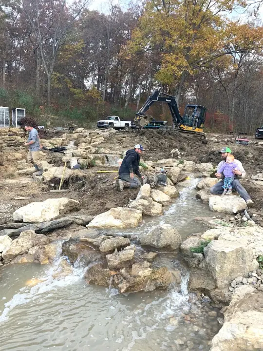 Several people placing large stones in a new stream bed in autumn, with an excavator behind
