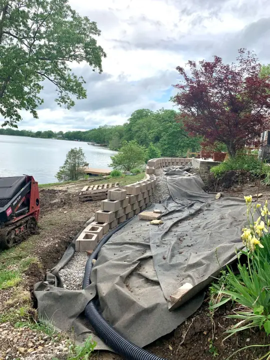 A wall of blocks, black fabric and a coiled drain pipe on a slope above a lake, with a compact track loader