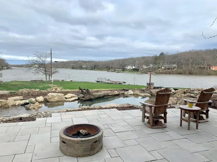 Light grey large-format paver patio with a round block fire pit and two wooden gliders, a boulder-edged pond below and the lake beyond