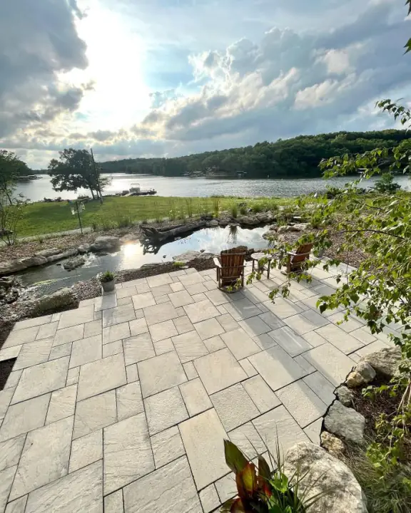 A paver patio in a random rectangular pattern overlooking a stone pond and a lake under a bright sky