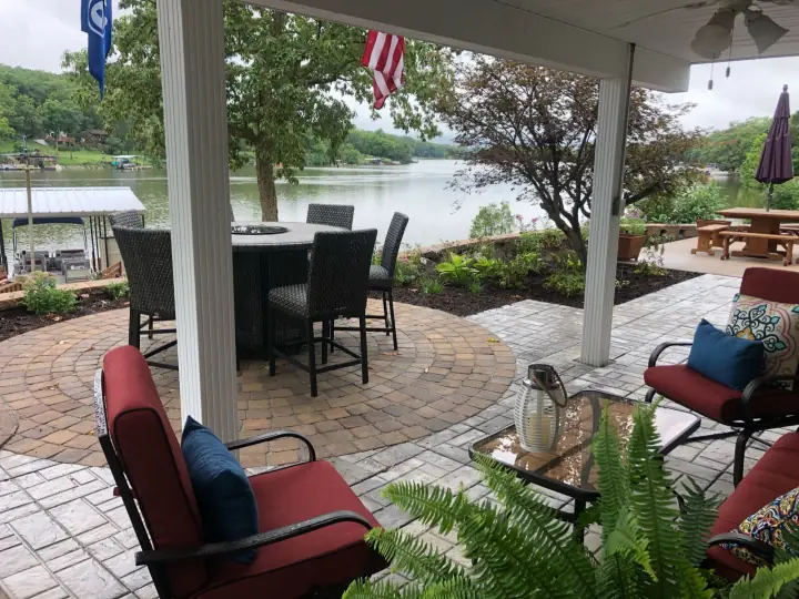 Outdoor dining set and red cushioned chairs on a paver circle under a covered porch, lake and boat dock beyond