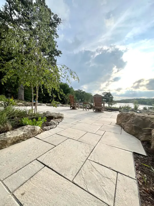 A long paver walkway of large rectangular stones running along a lake shore, edged with boulders and young trees