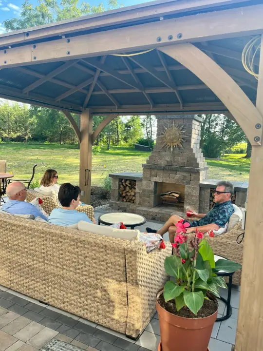 People relaxing on wicker seating under a wooden pavilion in front of an outdoor stone fireplace