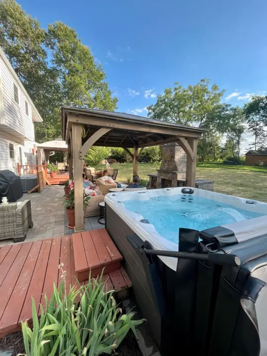 A wooden pavilion with a stone fireplace next to a hot tub on a deck and patio, with new plants
