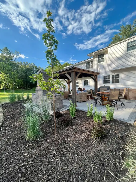 A wooden pavilion over a paver patio with outdoor furniture, new shrubs and grasses planted in mulch in front