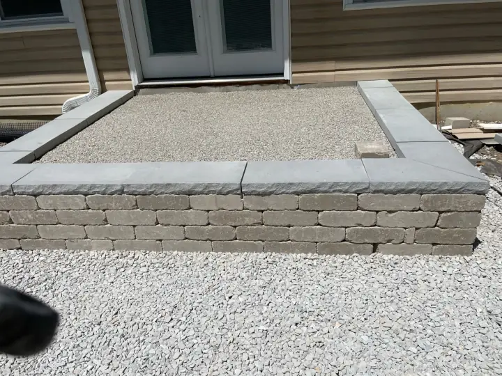 A raised patio border of tumbled wall block with smooth cap stones, filled with compacted gravel, beside a house door