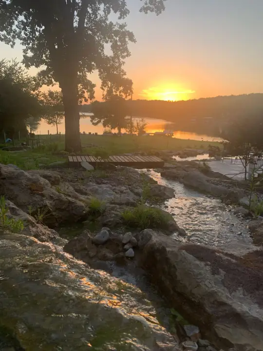 A stream running over rocks toward a lake at sunset, the sun low behind a tree