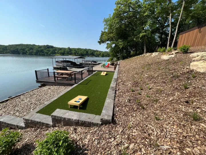 A strip of artificial turf with cornhole boards between a block wall and gravel, beside a lake and dock
